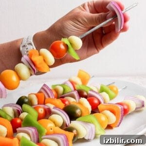 Rainbow Grilled Gnocchi Skewers with Roasted Veggies 6 A hand holds a metal skewer with pieces of onion, bell pepper, cherry tomatoes, and other vegetables above a plate of similar vegetable skewers.
