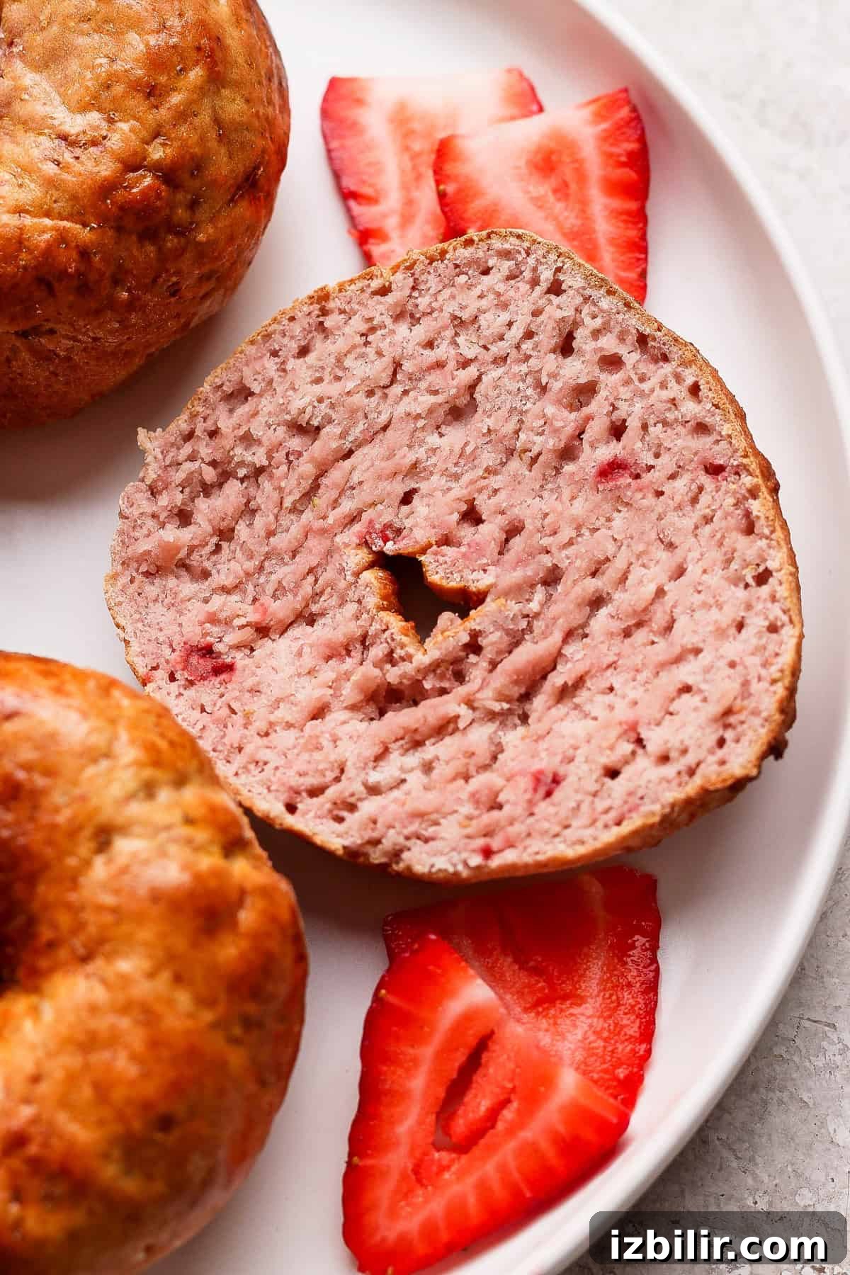 A sliced strawberry bagel on a plate with three fresh strawberry slices, next to a whole bagel.