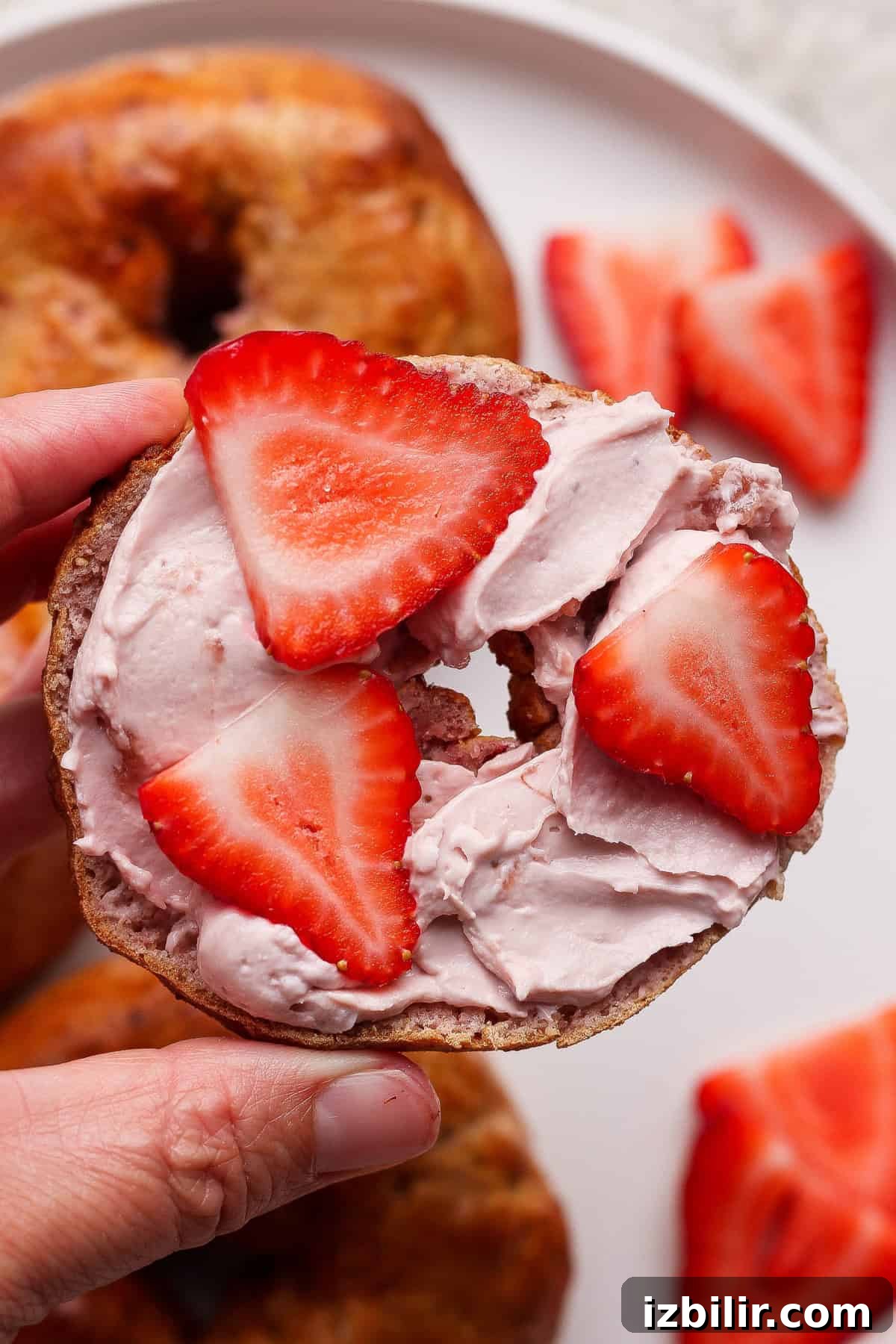A hand holds a bagel half topped with pink cream cheese and sliced strawberries, with more strawberries and bagels on a plate in the background.