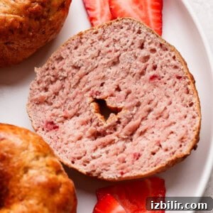 A sliced strawberry bagel is shown on a white plate with two fresh strawberry slices underneath.