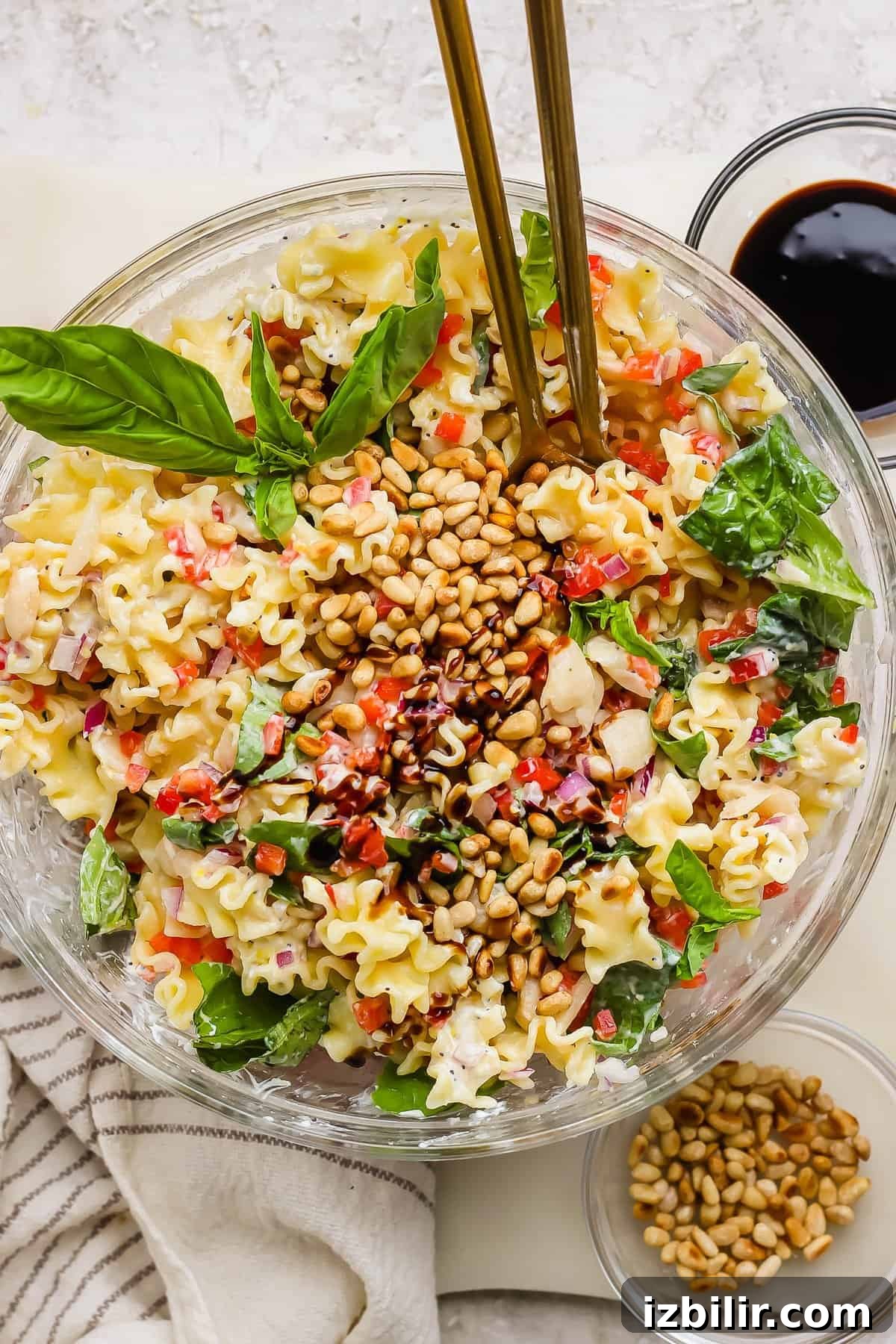 Creamy Cottage Cheese Pasta Salad Recipe 2 A glass bowl of pasta salad with spinach, pine nuts, red onion, and chopped vegetables, garnished with basil leaves and served with dressing on the side.