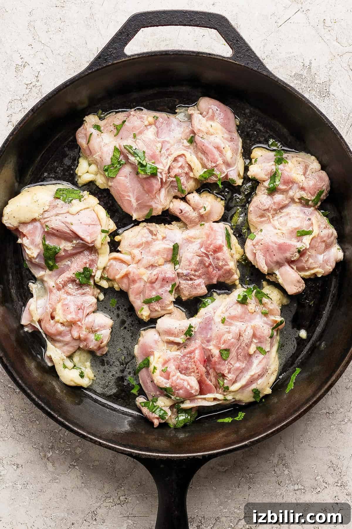Marinated chicken thighs ready to cook Raw marinated chicken pieces with herbs placed in a black cast iron skillet on a light countertop.
