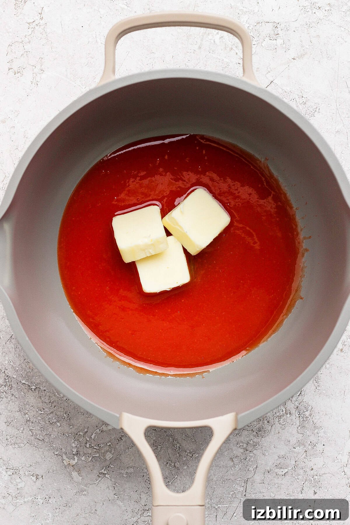 A grey pan contains a red liquid with three chunks of butter melting in it. The background is a light, textured surface.