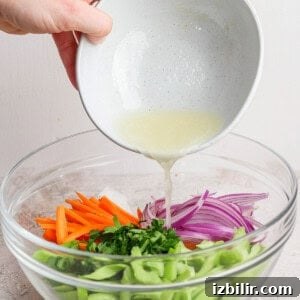 Person pouring a light dressing into a bowl of chopped vegetables including carrots, red onions, celery, and herbs.