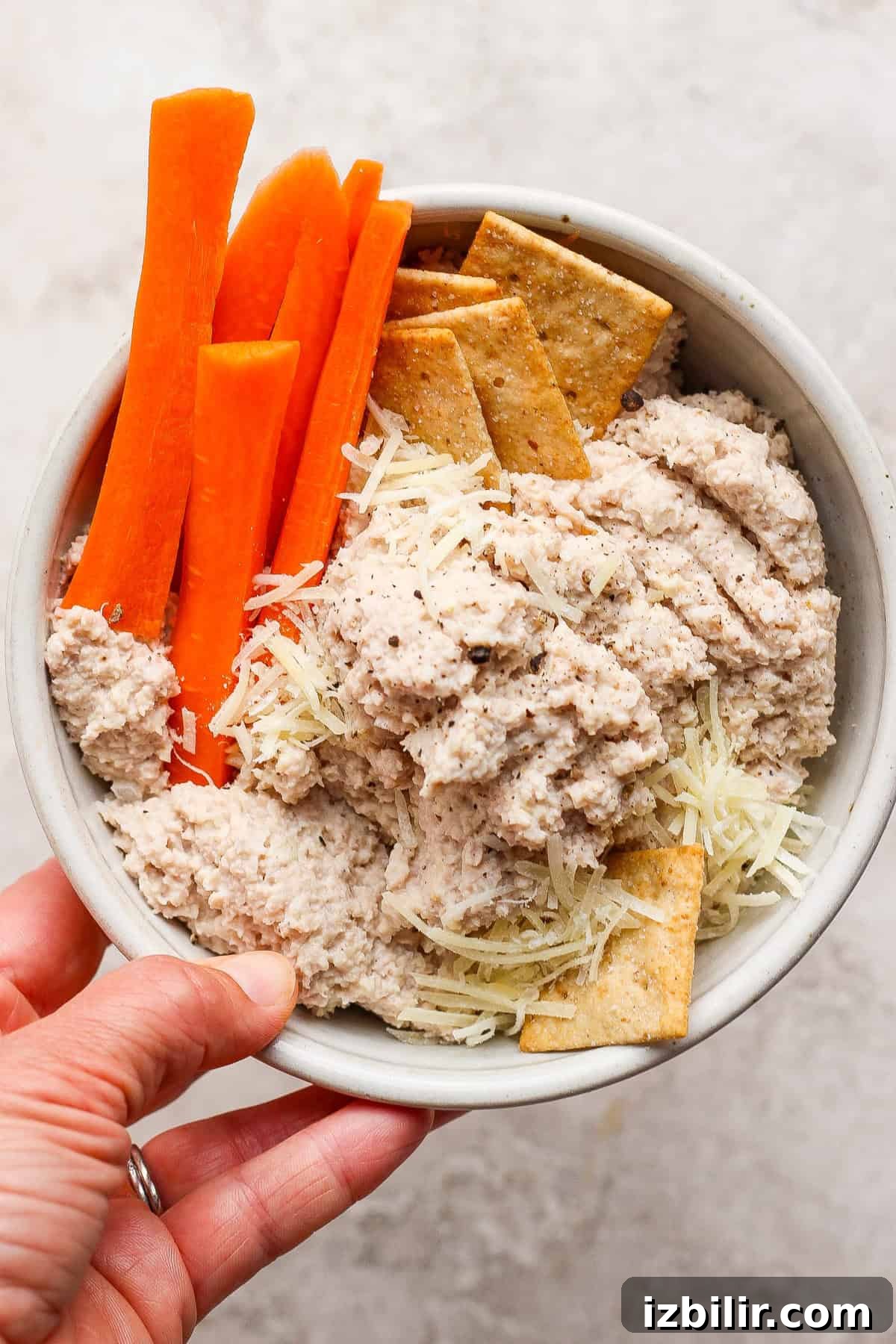 A hand holds a bowl containing carrot sticks, crackers, shredded cheese, and a creamy chicken caesar salad spread.