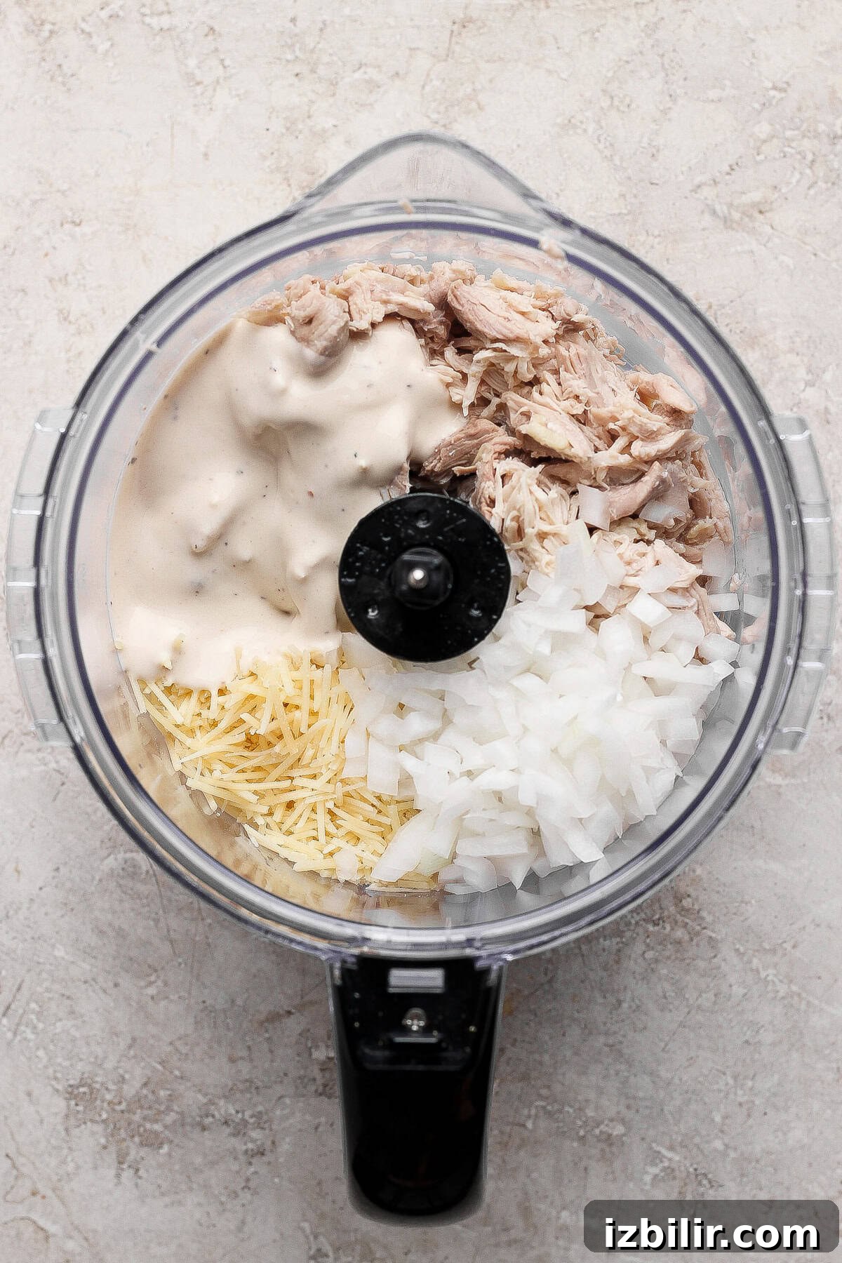 A food processor bowl containing shredded chicken, chopped onions, shredded cheese, and a creamy sauce is prepped for a flavorful chicken caesar salad spread, viewed from above.