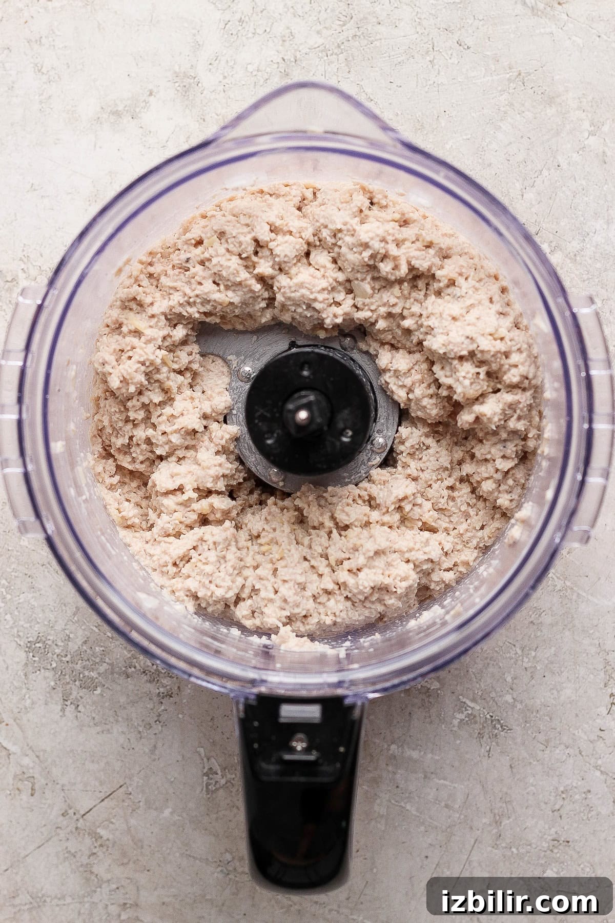 A food processor filled with a coarse, pale chicken caesar salad spread sits on a light-colored textured surface, viewed from above.