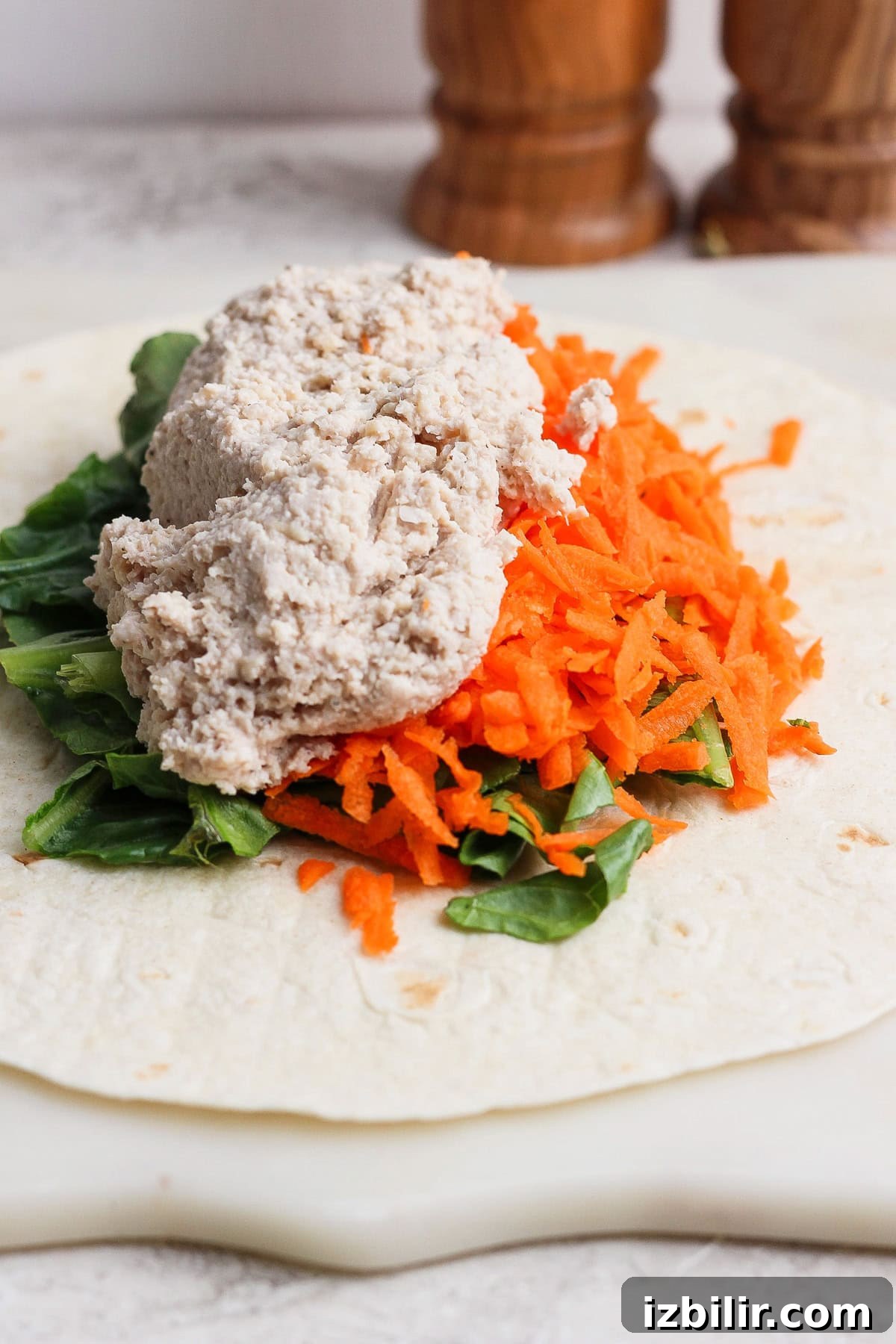 A flour tortilla topped with leafy greens, grated carrots, and a scoop of chicken caesar salad spread on a white surface.