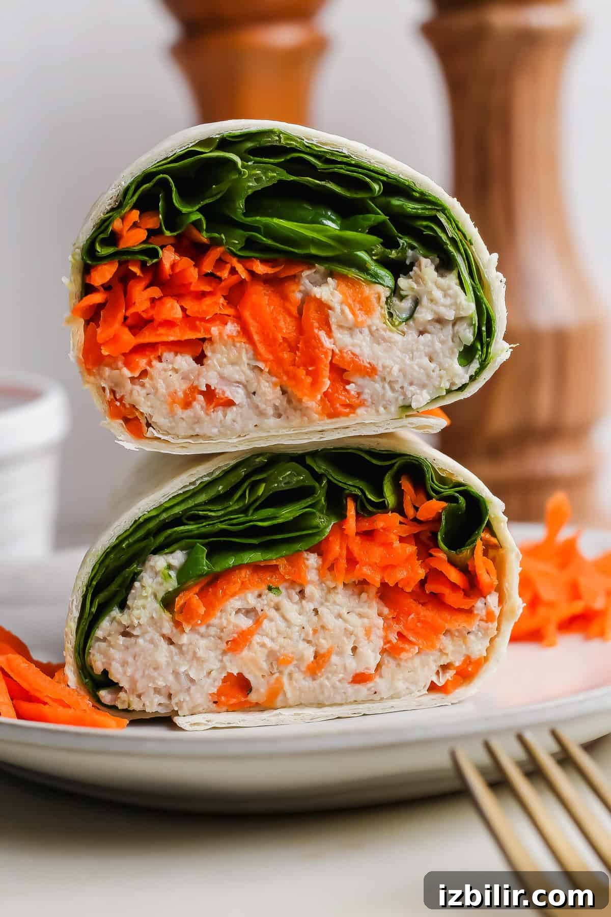 A halved wrap filled with shredded carrots, leafy greens, and a creamy chicken salad mixture, stacked on a white plate with a fork beside it.