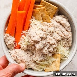 A bowl containing carrot sticks, crackers, shredded cheese, and a creamy dip, held in one hand.