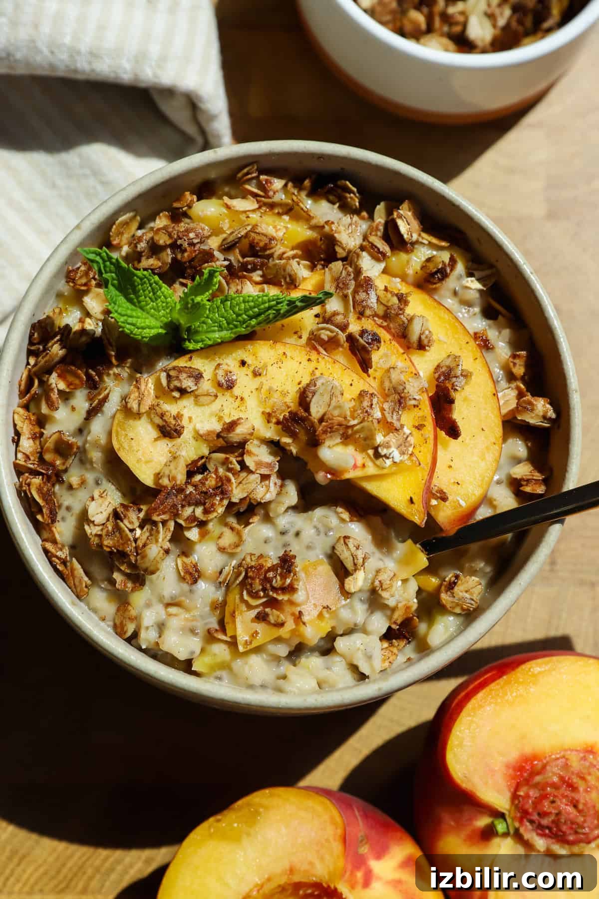 Baked Peach Cobbler Oatmeal Recipe 2 A bowl of oatmeal topped with granola, fresh peach slices, and a mint sprig, with a spoon inside and peach halves nearby on a wooden surface.
