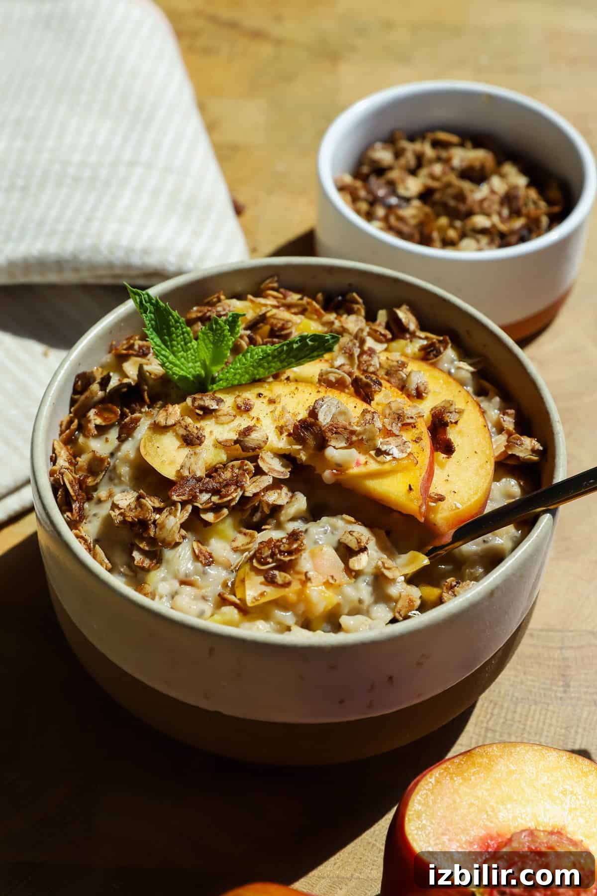 Baked Peach Cobbler Oatmeal Recipe 3 A bowl of oatmeal topped with peach slices, granola, and a mint sprig, with a spoon inside. A small bowl of granola and a peach half are nearby on a wooden surface.