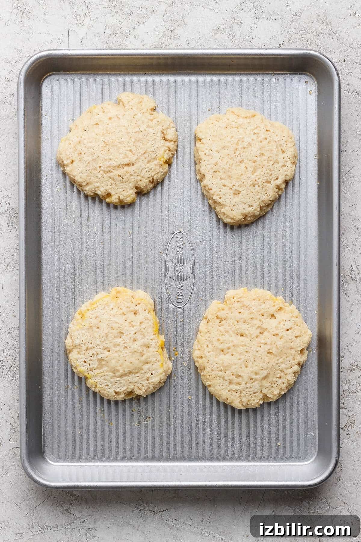 Four unbaked biscuit dough rounds are spaced evenly on a metal baking sheet placed on a light-colored surface.