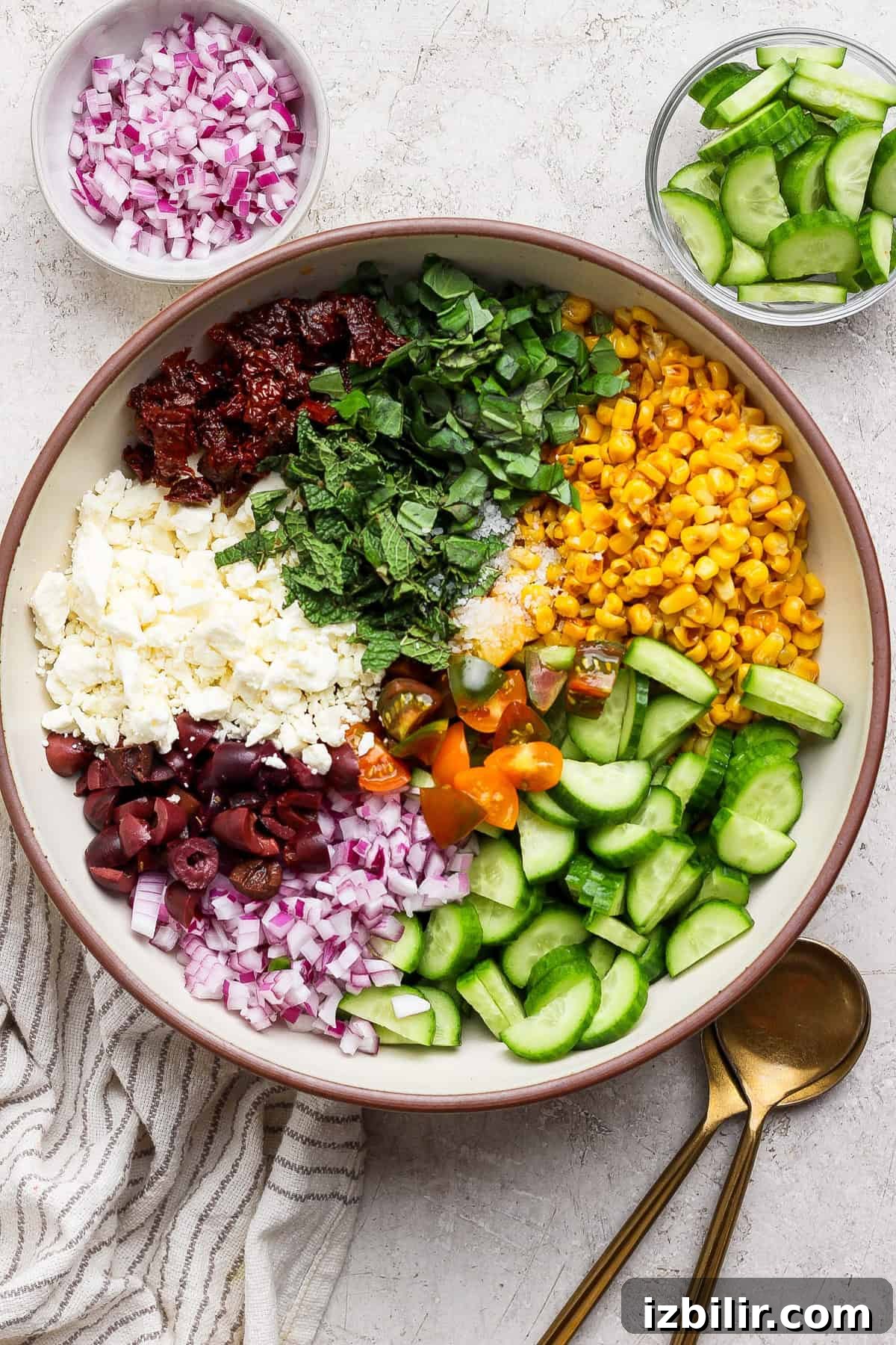 Mediterranean Corn Salad with Feta and Tomatoes 2 A large bowl containing chopped red onion, sun-dried tomatoes, feta cheese, olives, basil, mint, corn, cherry tomatoes, and cucumber, with bowls of onions and cucumbers on the side.