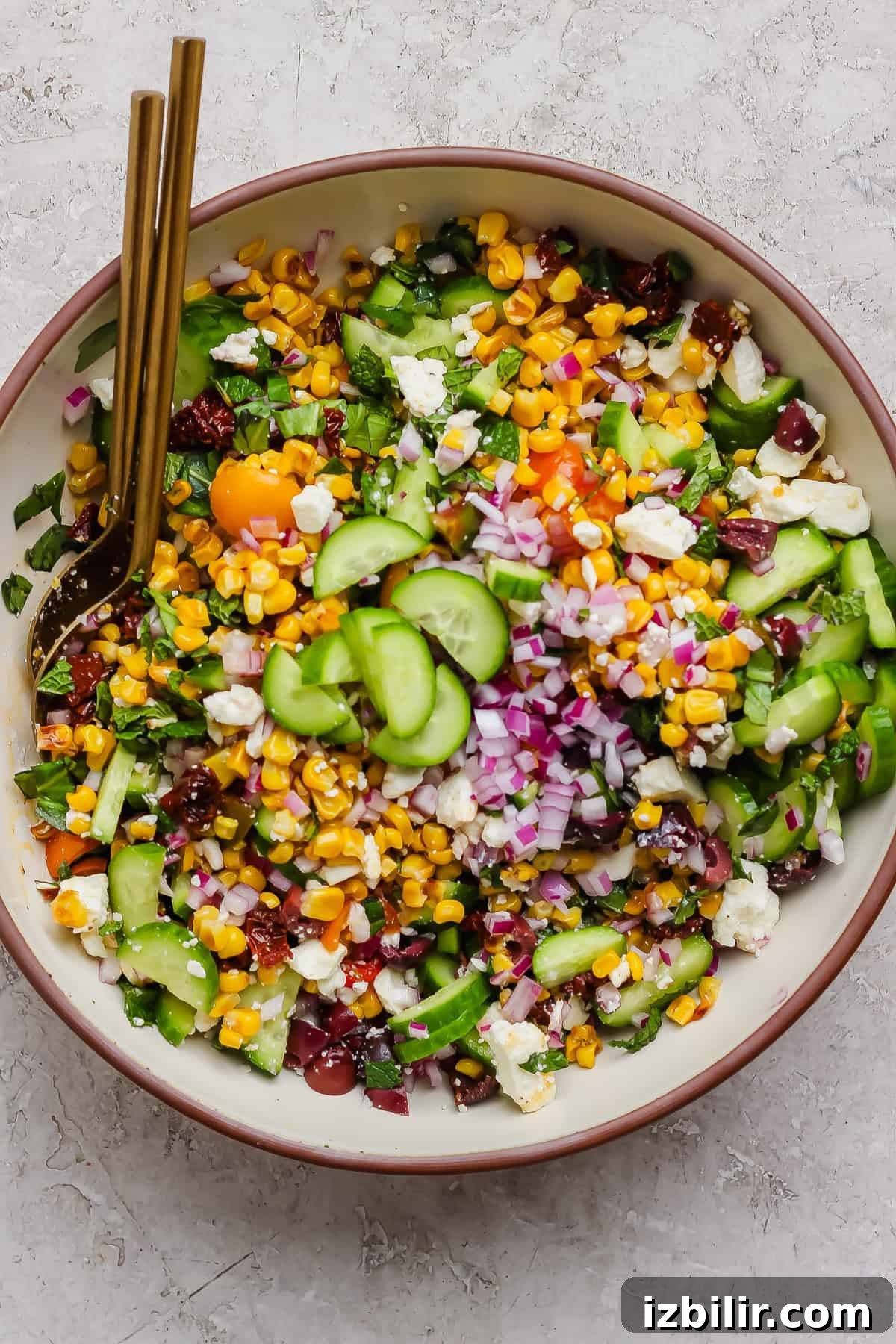 Mediterranean Corn Salad with Feta and Tomatoes 4 A bowl of Mediterranean corn salad with cucumbers, red onions, cherry tomatoes, feta cheese, leafy greens, and two gold forks.
