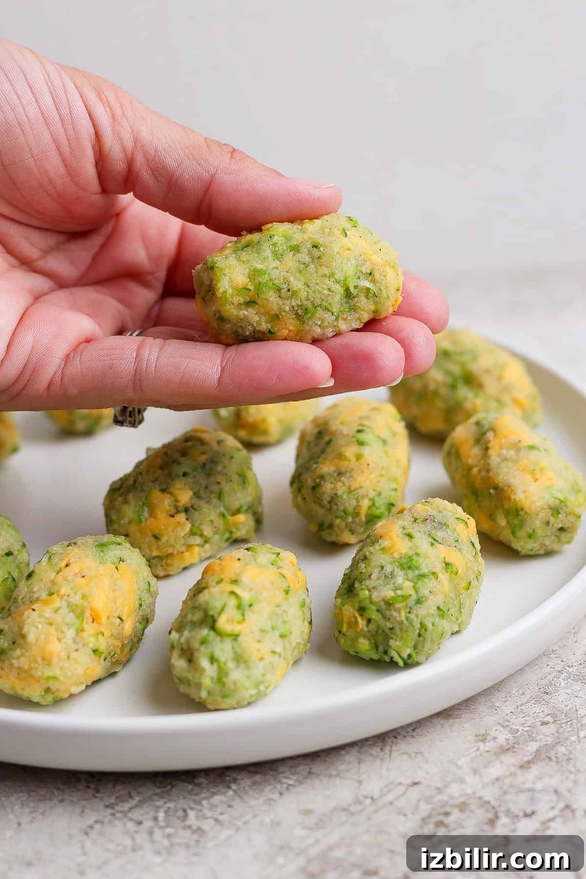 Crispy Air Fryer Zucchini Tots 5 A hand holds a homemade zucchini and cheese tot over a white plate with more tots arranged on it.