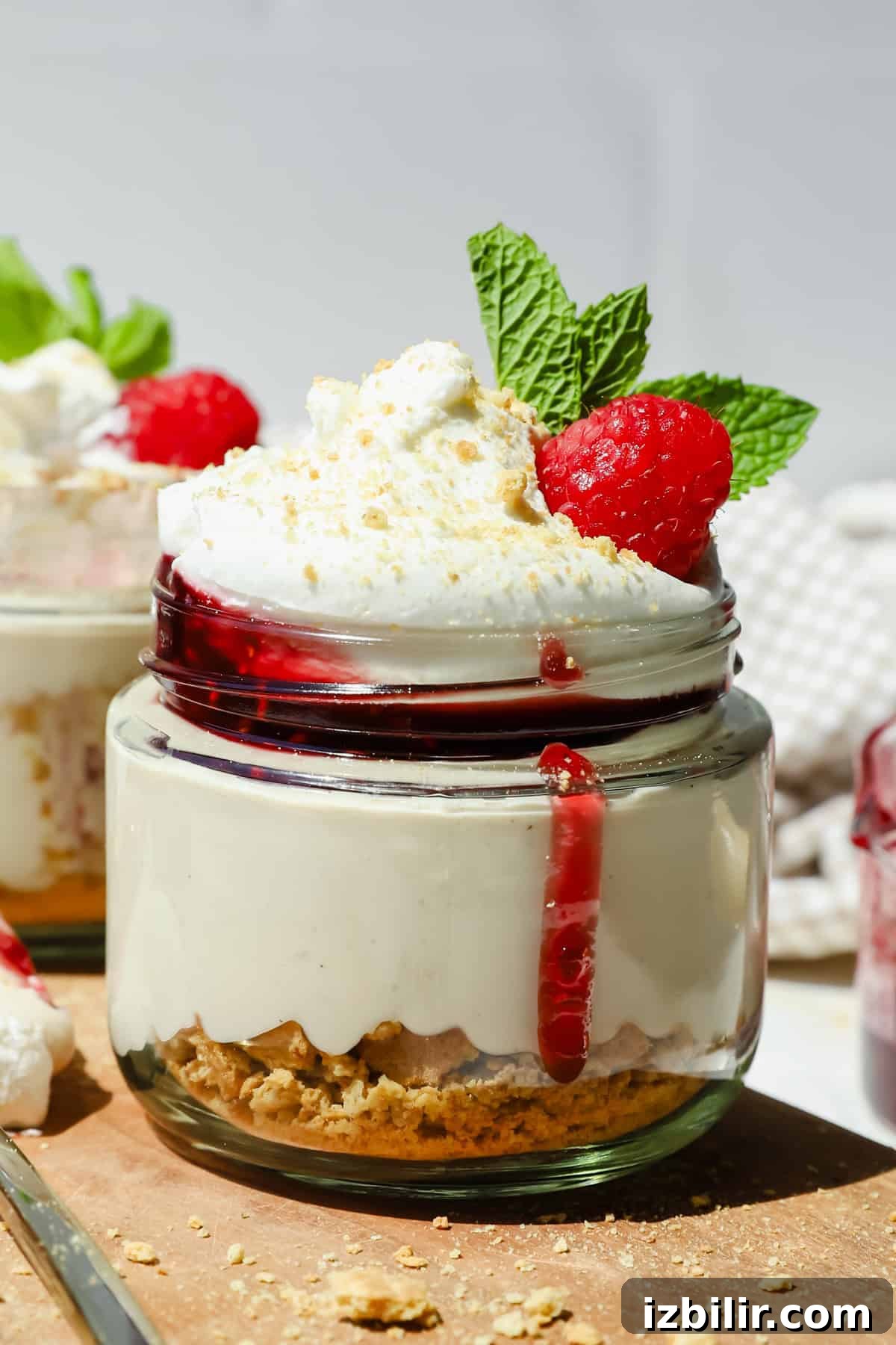 High-Protein Cheesecake Jar Recipe with Berries 2 A small glass jar of cheesecake topped with whipped cream, raspberry sauce, a fresh raspberry, and mint leaves, with a layer of crushed biscuits at the bottom.