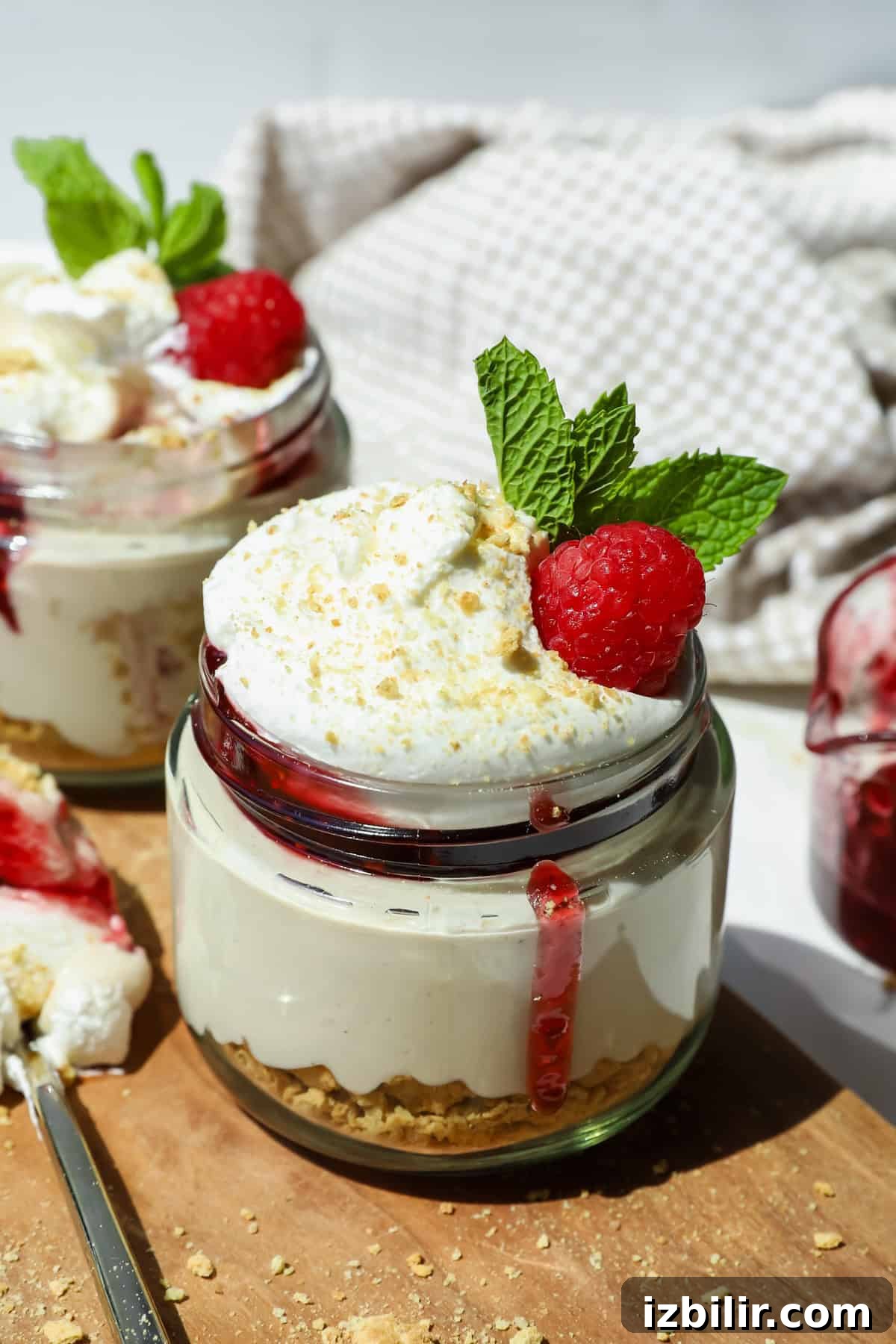 High-Protein Cheesecake Jar Recipe with Berries 5 A glass jar filled with layered cheesecake, graham cracker crust, whipped cream, a raspberry, and mint leaves, with berry sauce dripping down the side.