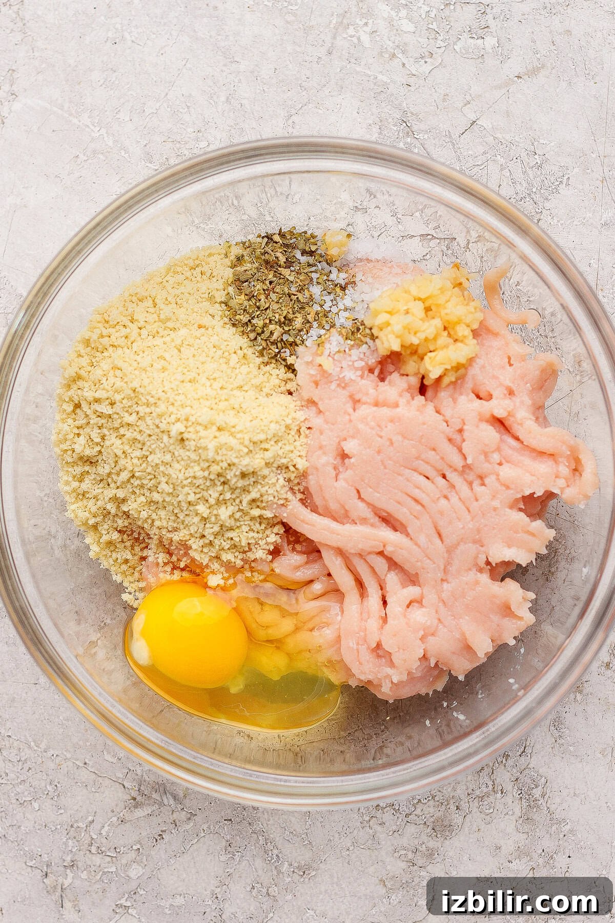 Skillet Chicken Meatballs with Summer Squash and Orzo 3 A glass bowl containing ground chicken, a cracked egg, breadcrumbs, minced garlic, salt, and dried herbs on a light textured surface.