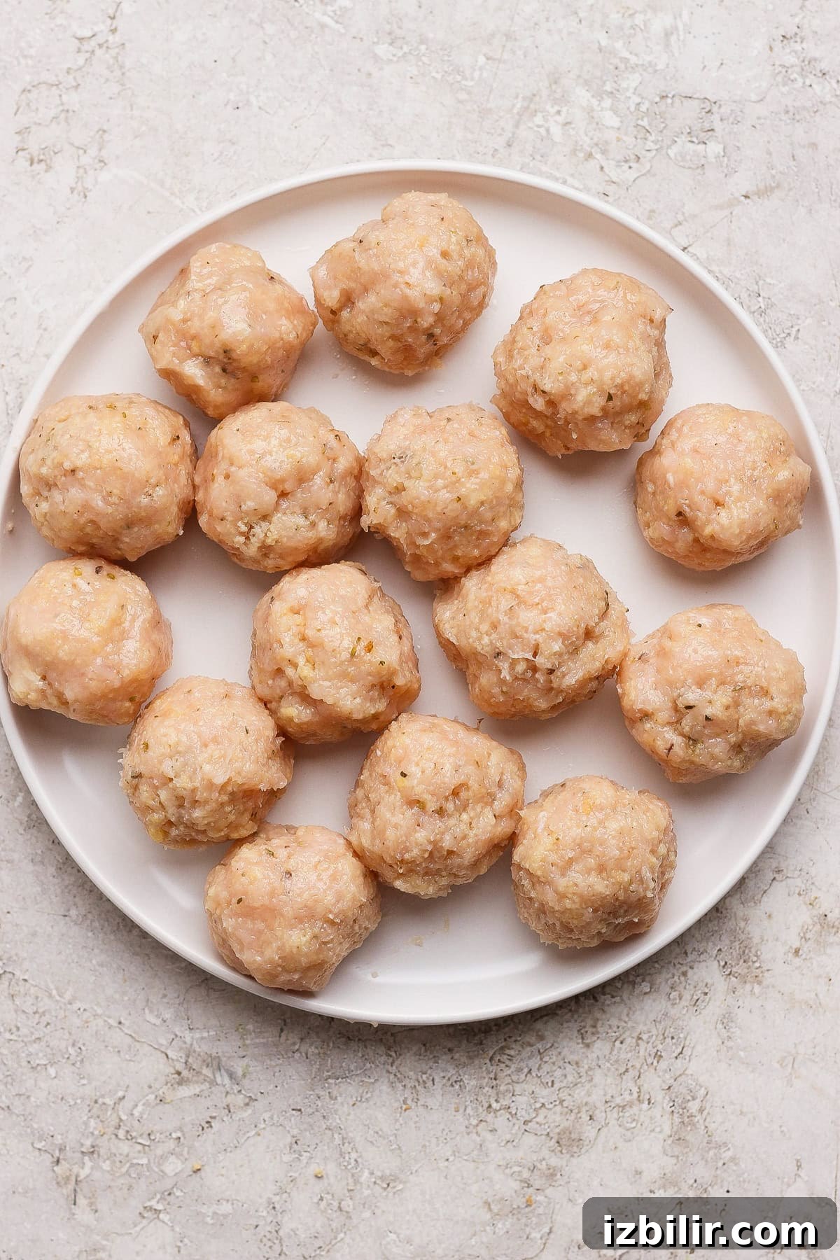 Skillet Chicken Meatballs with Summer Squash and Orzo 4 A white plate with sixteen raw, uncooked meatballs arranged in a circular pattern on a light textured surface.