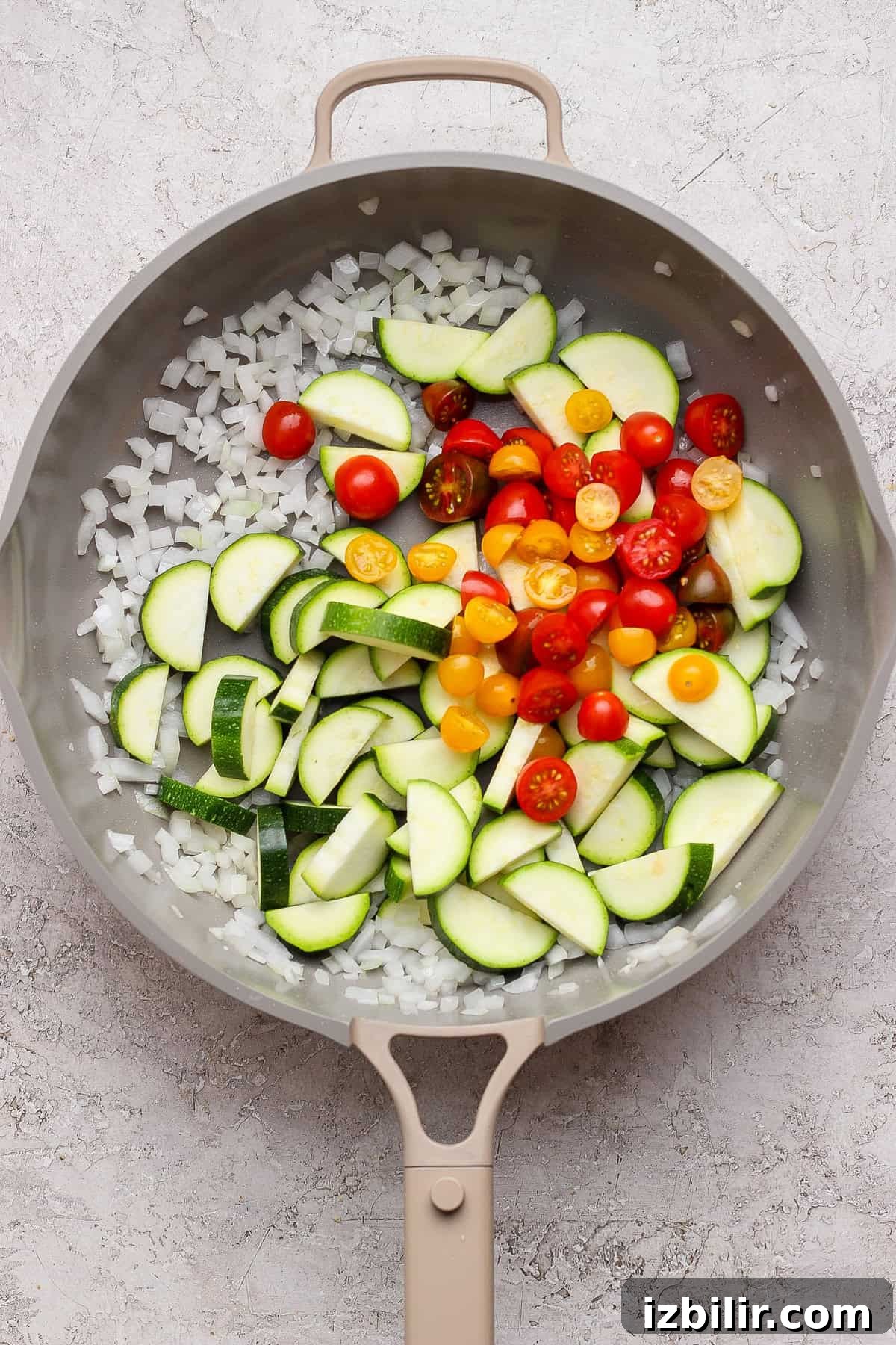 Skillet Chicken Meatballs with Summer Squash and Orzo 5 A frying pan contains chopped onions, sliced zucchini, and halved cherry tomatoes on a light textured surface.