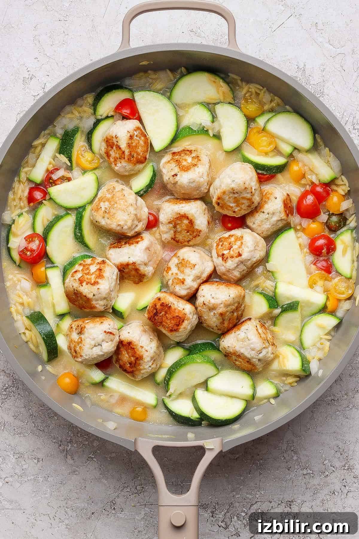 Skillet Chicken Meatballs with Summer Squash and Orzo 6 A skillet with browned meatballs, sliced zucchini, yellow squash, cherry tomatoes, and orzo in a light sauce on a gray surface.