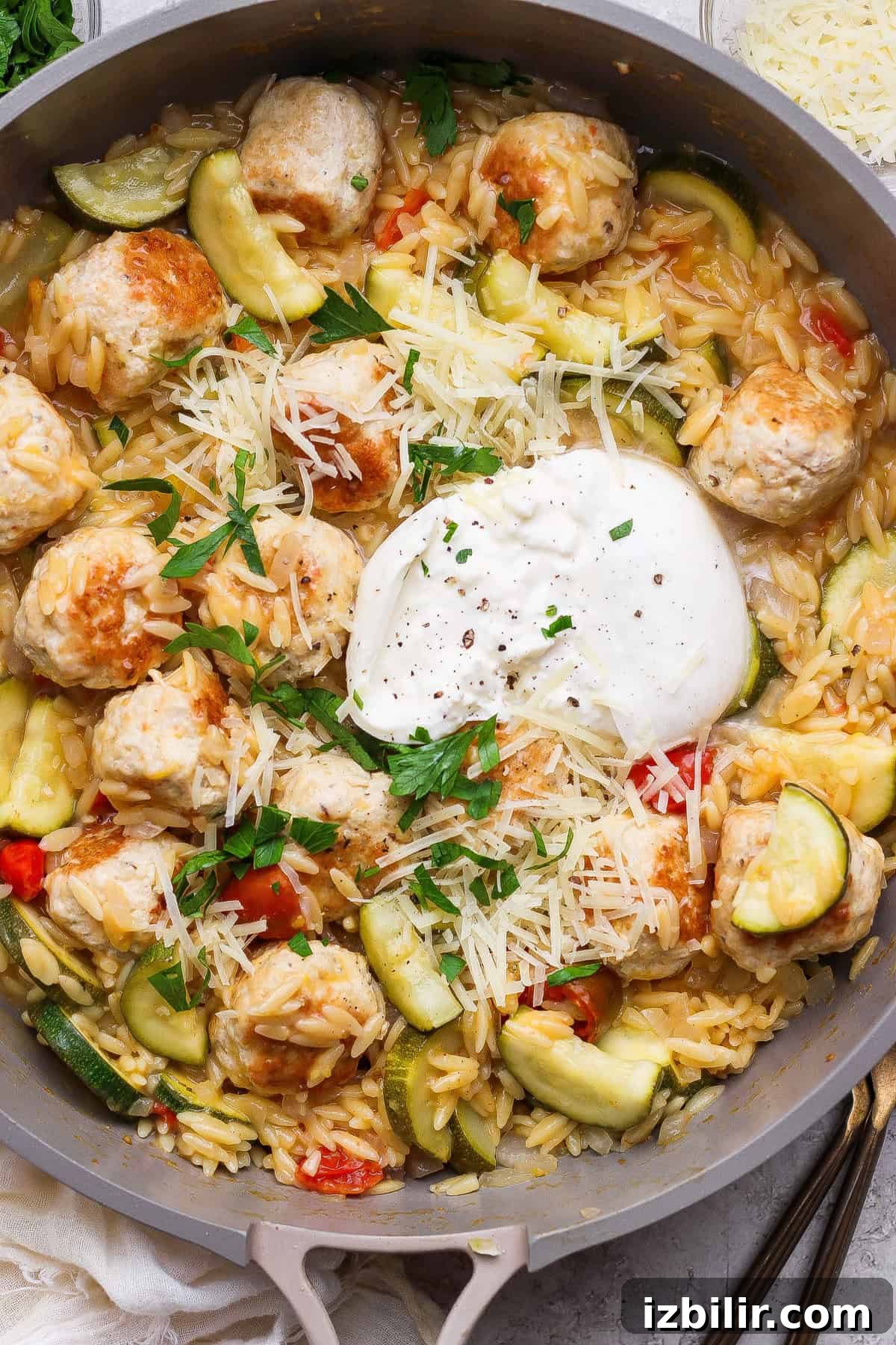 Skillet Chicken Meatballs with Summer Squash and Orzo 7 A skillet filled with orzo pasta, chicken meatballs, sliced zucchini, red peppers, grated cheese, fresh parsley, and a dollop of burrata cheese in the center.