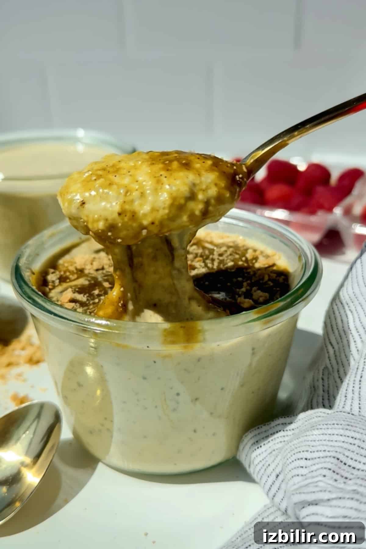 Creamy Blended Pistachio Overnight Oats Recipe 2 A spoon lifts creamy overnight oats from a glass jar, topped with a drizzle of syrup and a sprinkle of crumbs, with raspberries and a striped cloth nearby.