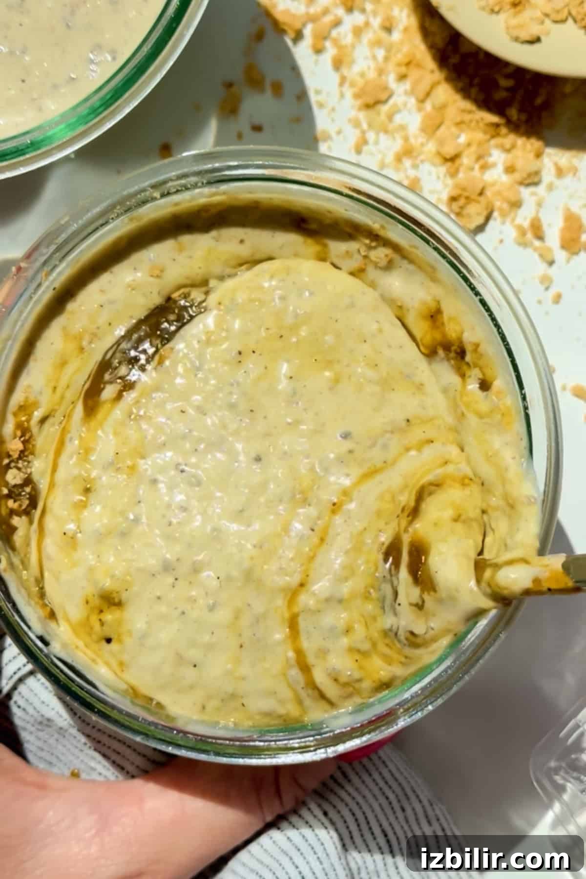 Creamy Blended Pistachio Overnight Oats Recipe 4 A glass bowl containing a creamy, beige mixture with swirls and specks, being stirred with a spoon. Crumbled pieces are scattered nearby on a white surface.