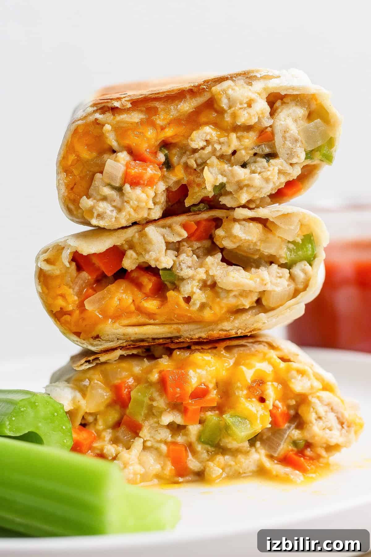 High-Protein Buffalo Chicken Burritos 2 Three halves of a burrito filled with scrambled eggs, cheese, and diced vegetables are stacked on a plate next to celery sticks.