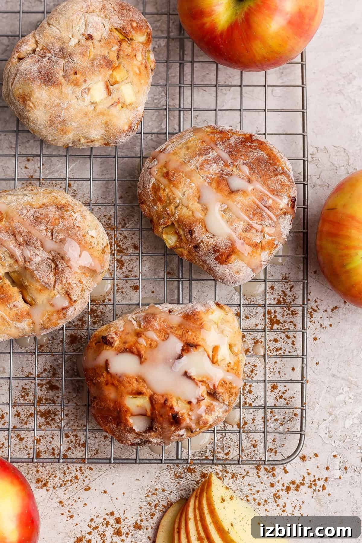 Crispy Air Fryer Apple Fritters with Glaze 5 Four apple-studded pastries with glaze sit on a cooling rack, surrounded by apples, cinnamon, and apple slices on a light surface.