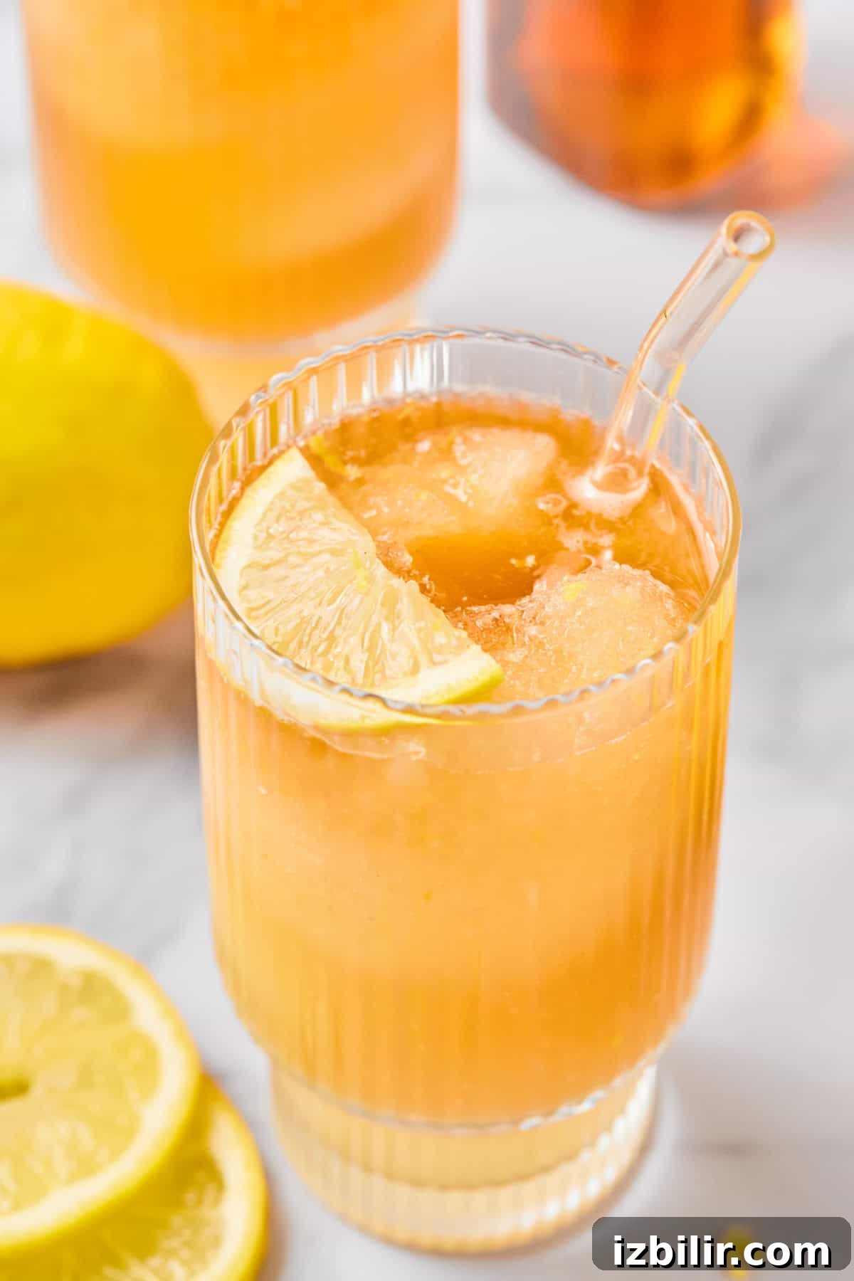 A glass of iced lemon drink with a slice of lemon on the rim and a glass straw, placed on a light surface with lemon slices nearby.