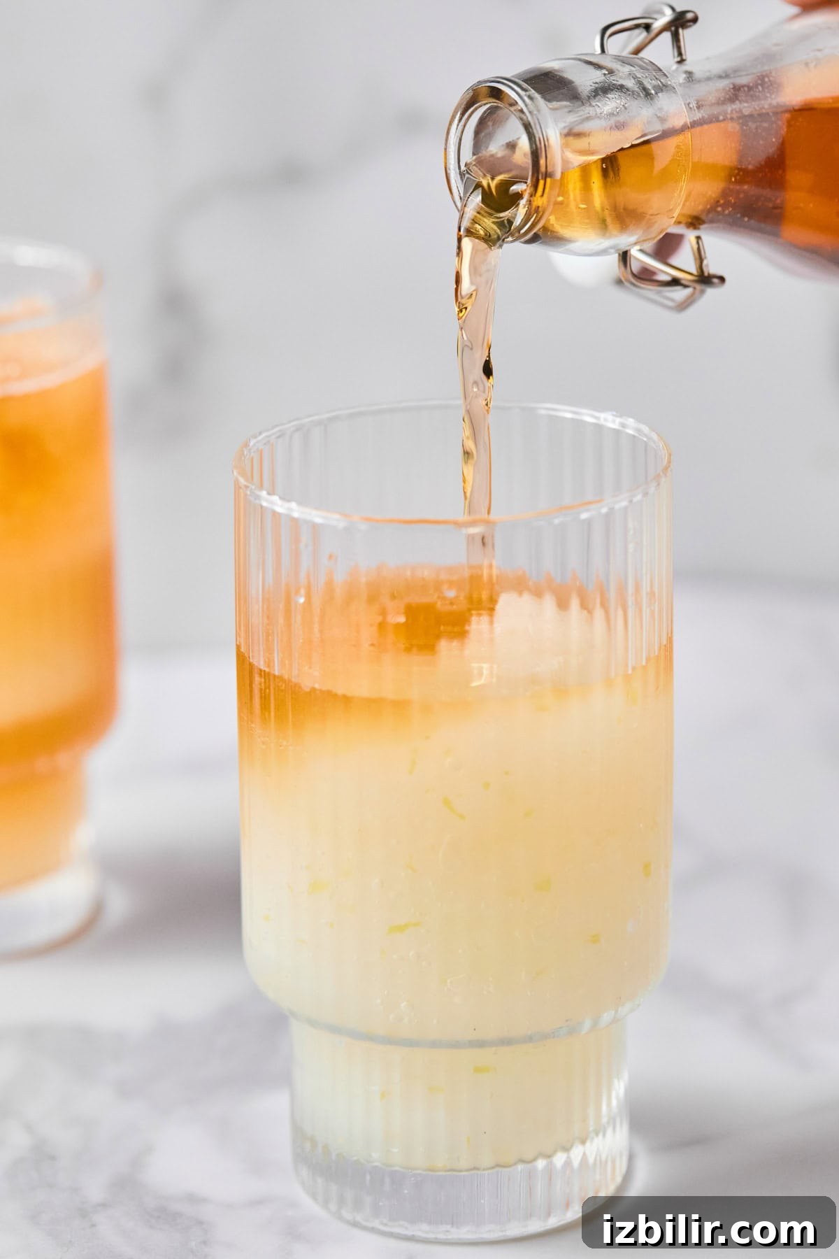 A glass with a pale yellow liquid and ice is being filled with an orange liquid from a glass bottle. Another similar drink is in the background.