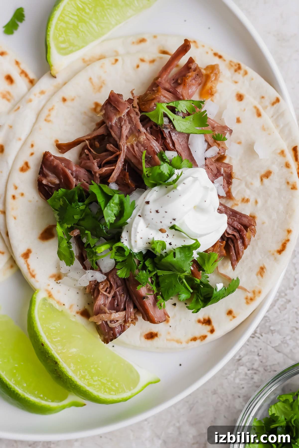 A soft flour tortilla topped with shredded meat, chopped onions, cilantro, and a dollop of sour cream, served with lime wedges on a white plate.