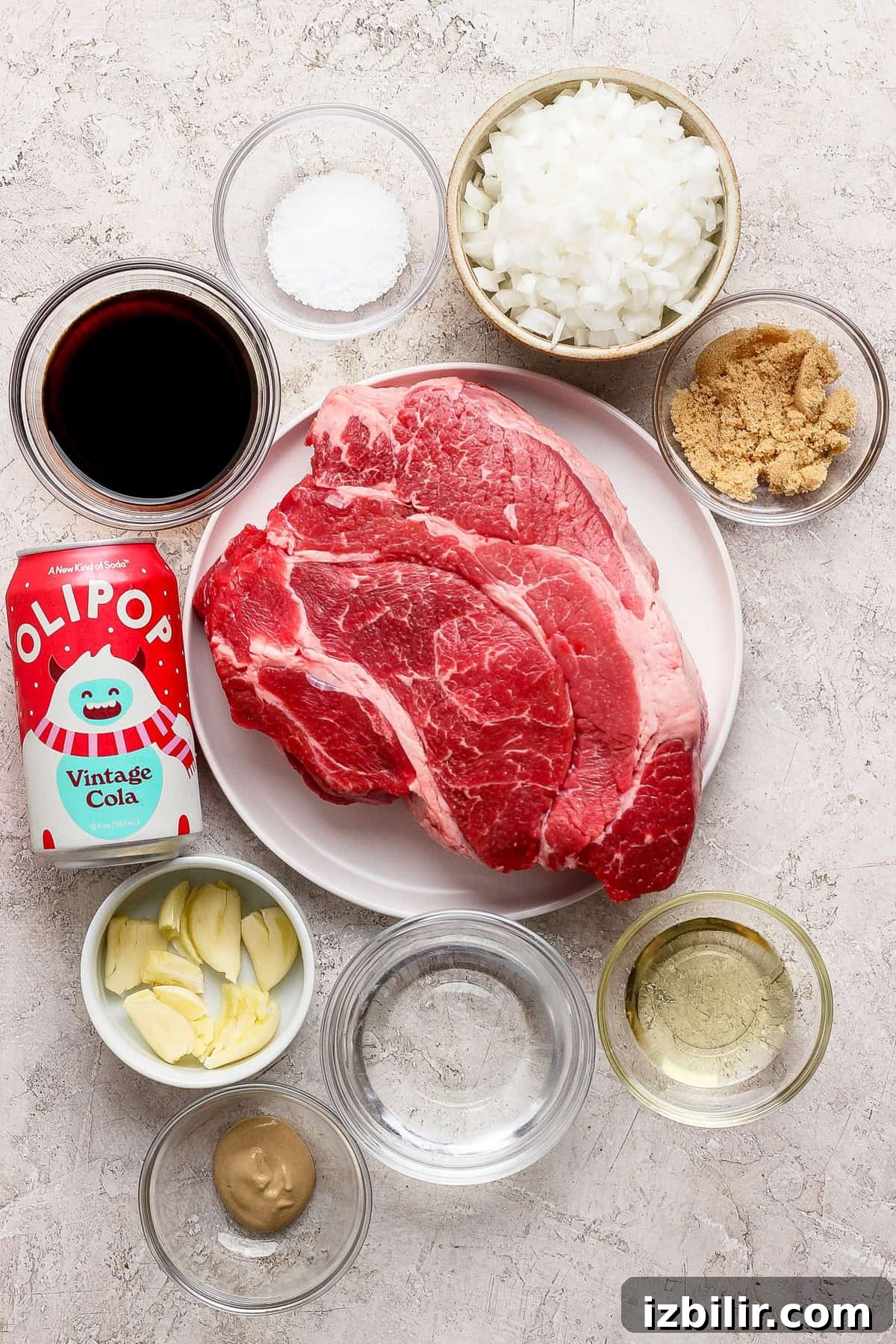 Raw chuck roast on a plate, surrounded by bowls of chopped onions, brown sugar, garlic, salt, vinegar, mustard, oil, soy sauce, and a can of OLIPOP Vintage Cola on a light surface.