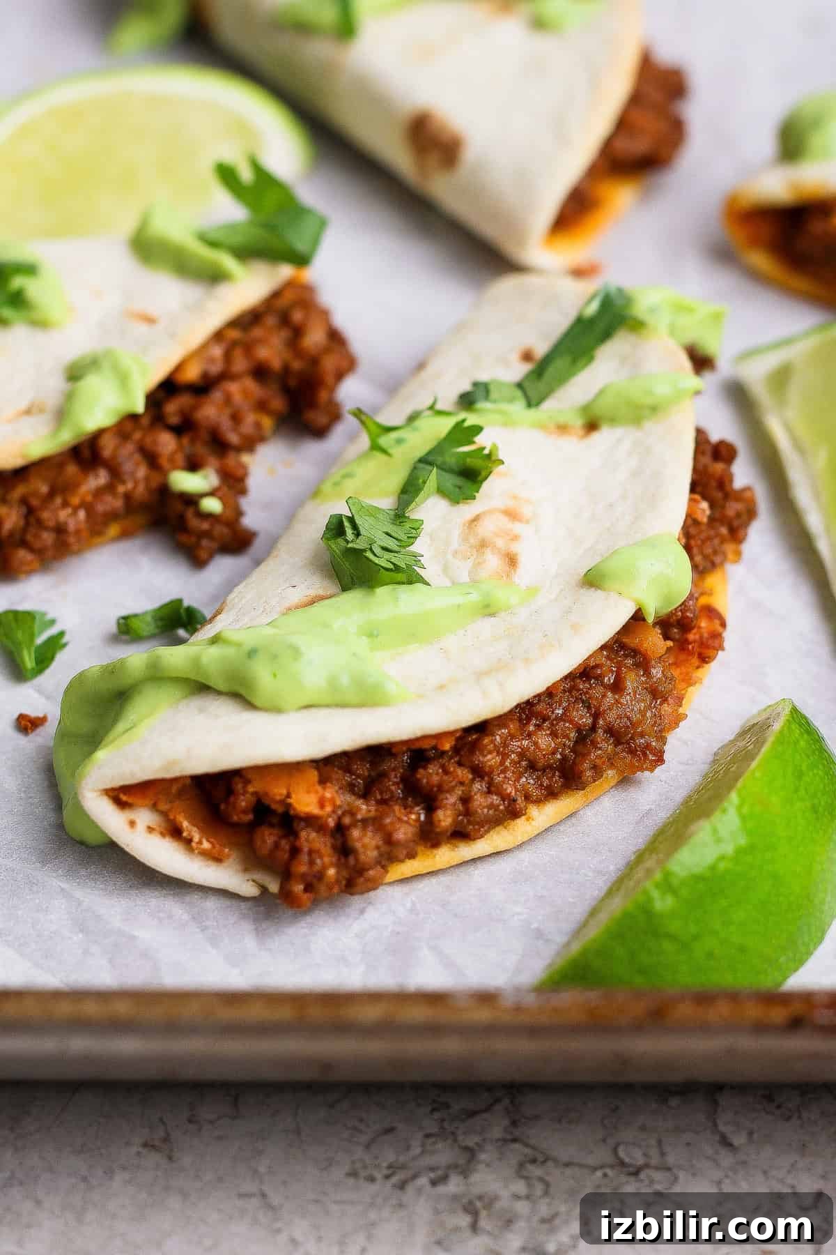 Sheet Pan Tacos with Creamy Avocado Crema Recipe 2 A taco filled with seasoned ground meat, topped with green sauce and cilantro, served on a tray with lime wedges.