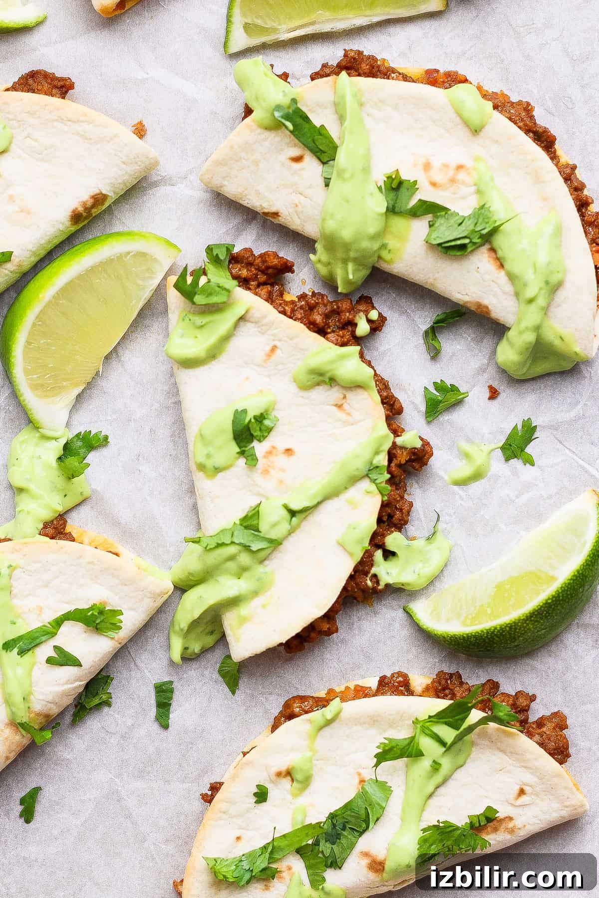 Sheet Pan Tacos with Creamy Avocado Crema Recipe 4 Three folded tacos filled with ground beef, topped with green sauce and fresh cilantro, are arranged on parchment paper with lime wedges.