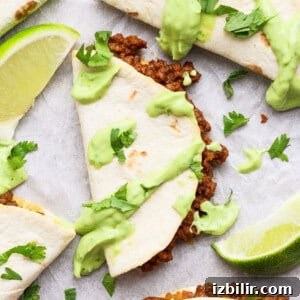 Sheet Pan Tacos with Creamy Avocado Crema Recipe 5 Taco halves with ground meat filling, topped with green sauce and cilantro, served with lime wedges on a white surface.