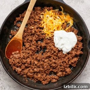 Sheet Pan Tacos with Creamy Avocado Crema Recipe 6 Ground beef in a cast iron skillet with shredded cheese and a dollop of Greek yogurt, being stirred with a wooden spoon.