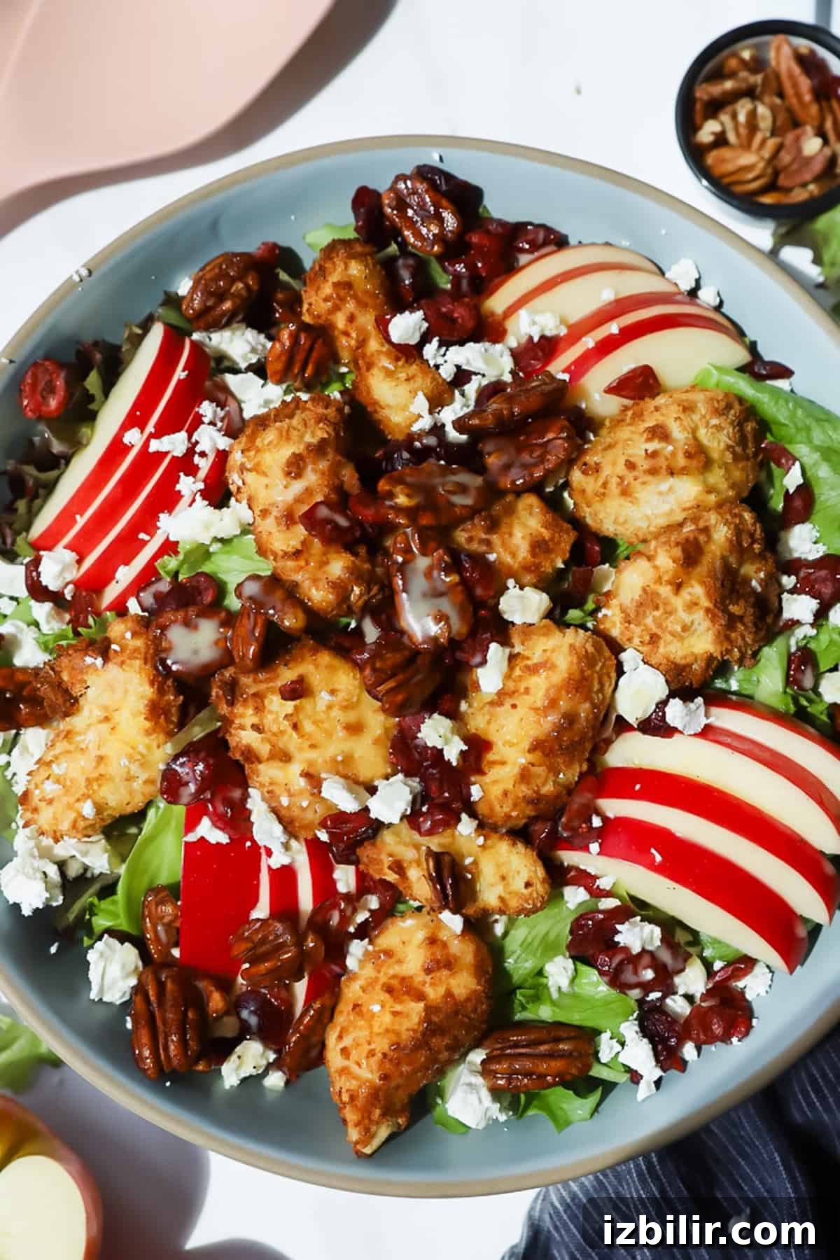 A salad with breaded chicken pieces, sliced red apples, pecans, dried cranberries, lettuce, and crumbled cheese, drizzled with dressing in a gray bowl.