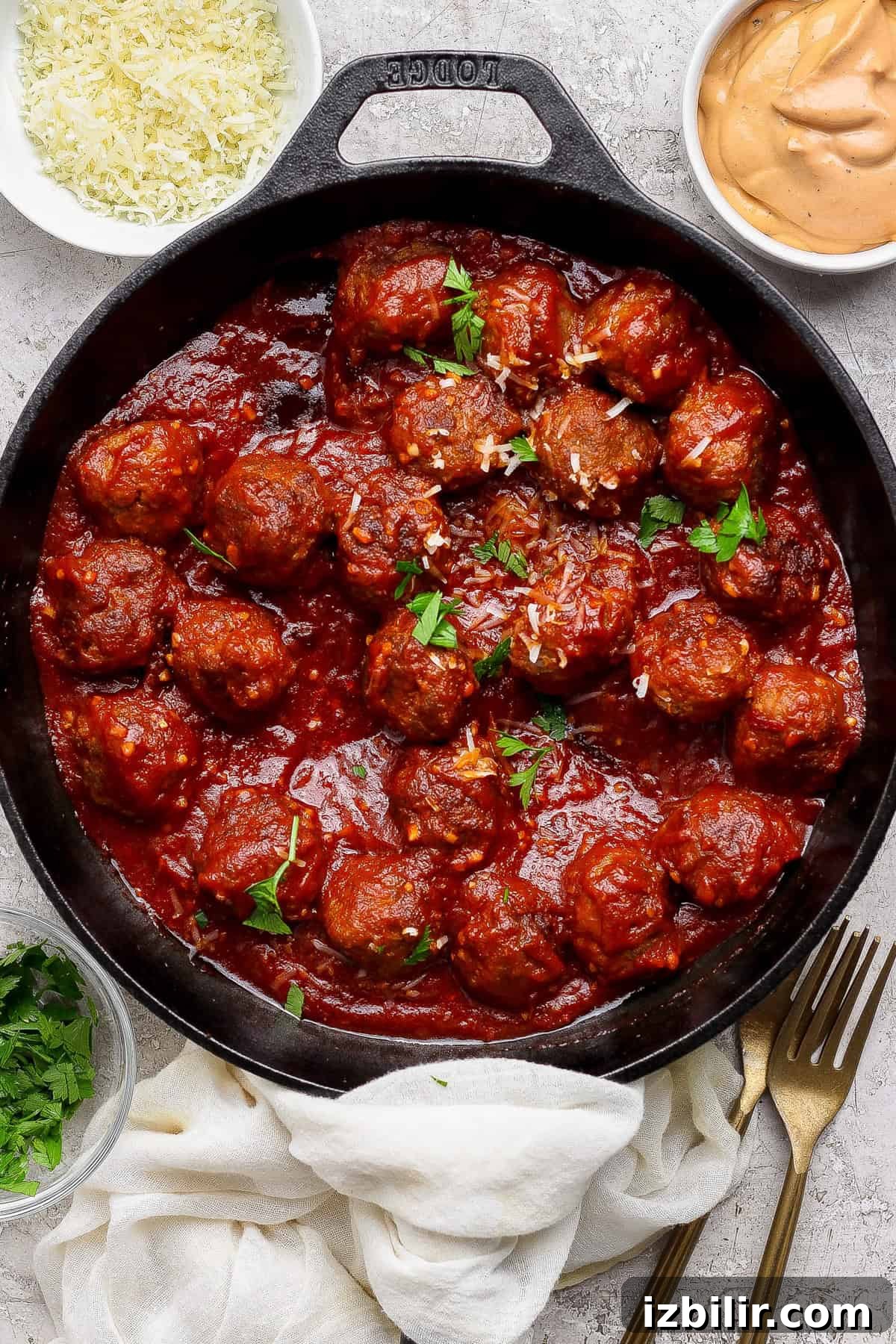 A cast iron skillet filled with meatballs in tomato sauce, garnished with parsley, surrounded by bowls of grated cheese, sauce, and chopped herbs.