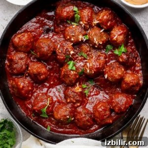 A cast iron skillet filled with meatballs in tomato sauce, garnished with parsley, surrounded by bowls of grated cheese, sauce, and chopped herbs.