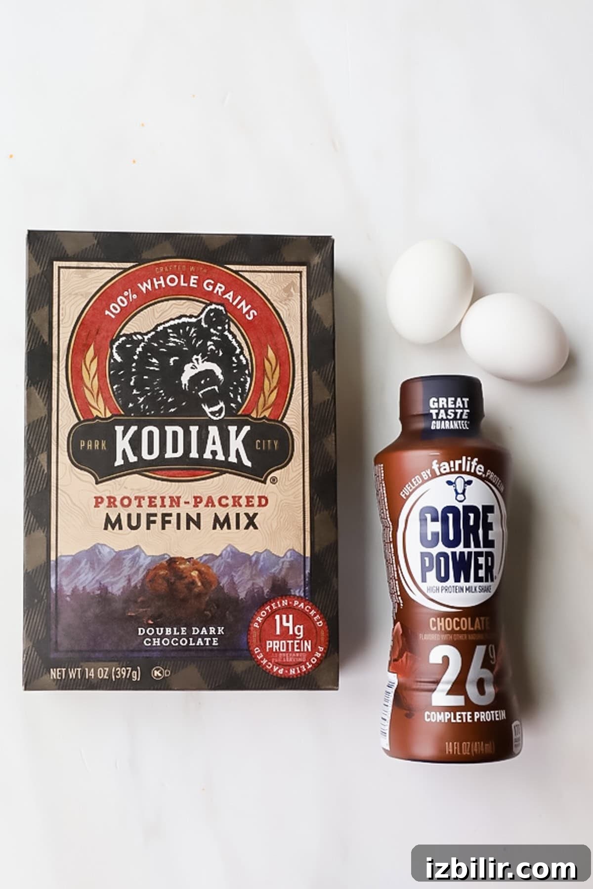 Chocolate Kodiak Protein Muffins Recipe 3 A box of Kodiak protein-packed muffin mix, a bottle of Core Power chocolate protein shake, and three white eggs are arranged on a white surface.