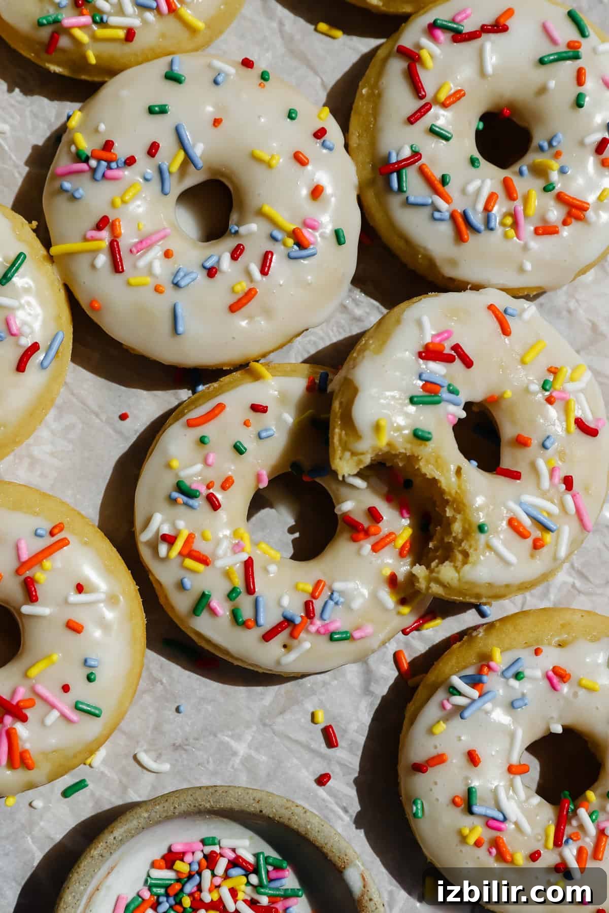 Glazed donuts topped with colorful sprinkles are arranged on parchment paper, with one donut partially eaten and a small bowl of extra sprinkles nearby.
