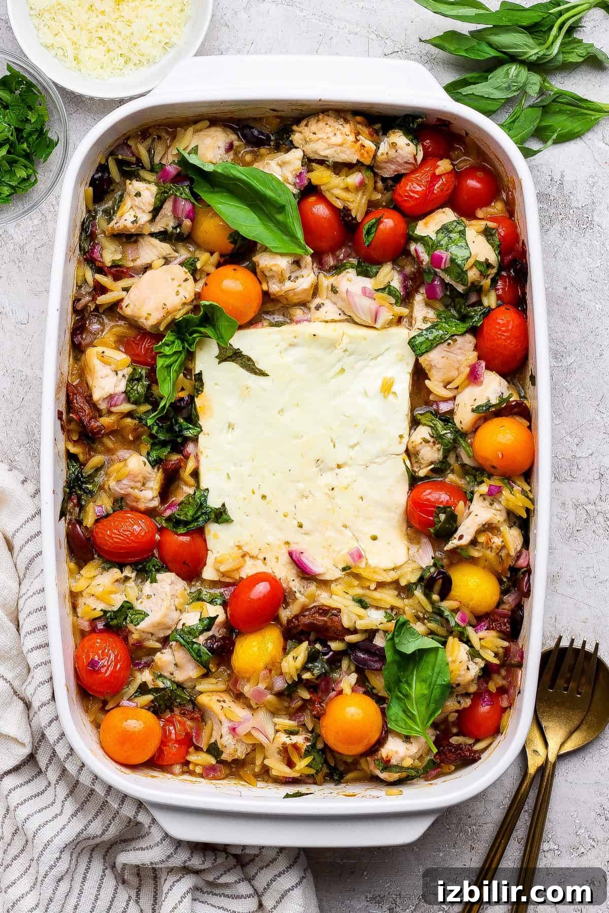 A baked casserole dish filled with chicken, cherry tomatoes, orzo pasta, red onions, and a large block of feta cheese in the center, garnished with fresh basil.