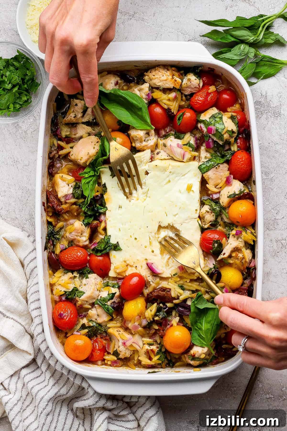 A person uses two forks to serve a baked dish with chicken, cherry tomatoes, greens, orzo, and feta cheese in a white casserole dish. Fresh herbs are visible on the side.
