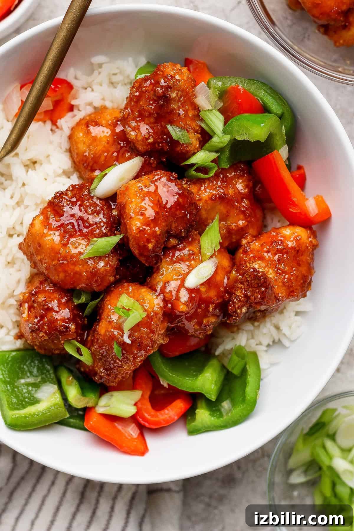 A bowl of white rice topped with glazed chicken pieces, chopped green onions, and sliced red and green bell peppers.