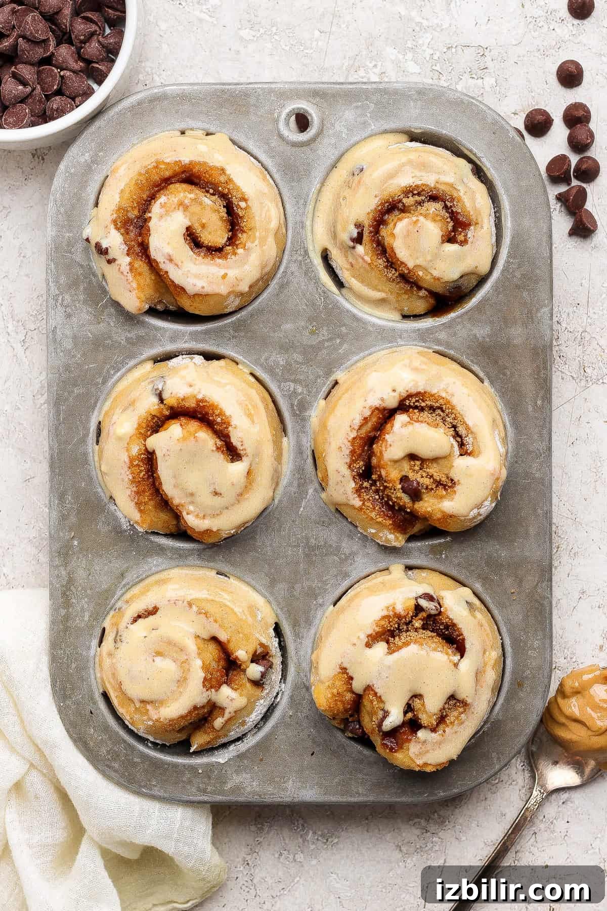 Six unbaked cinnamon rolls with chocolate chips in a metal muffin tin, with a bowl of chocolate chips and a spoon with peanut butter nearby.