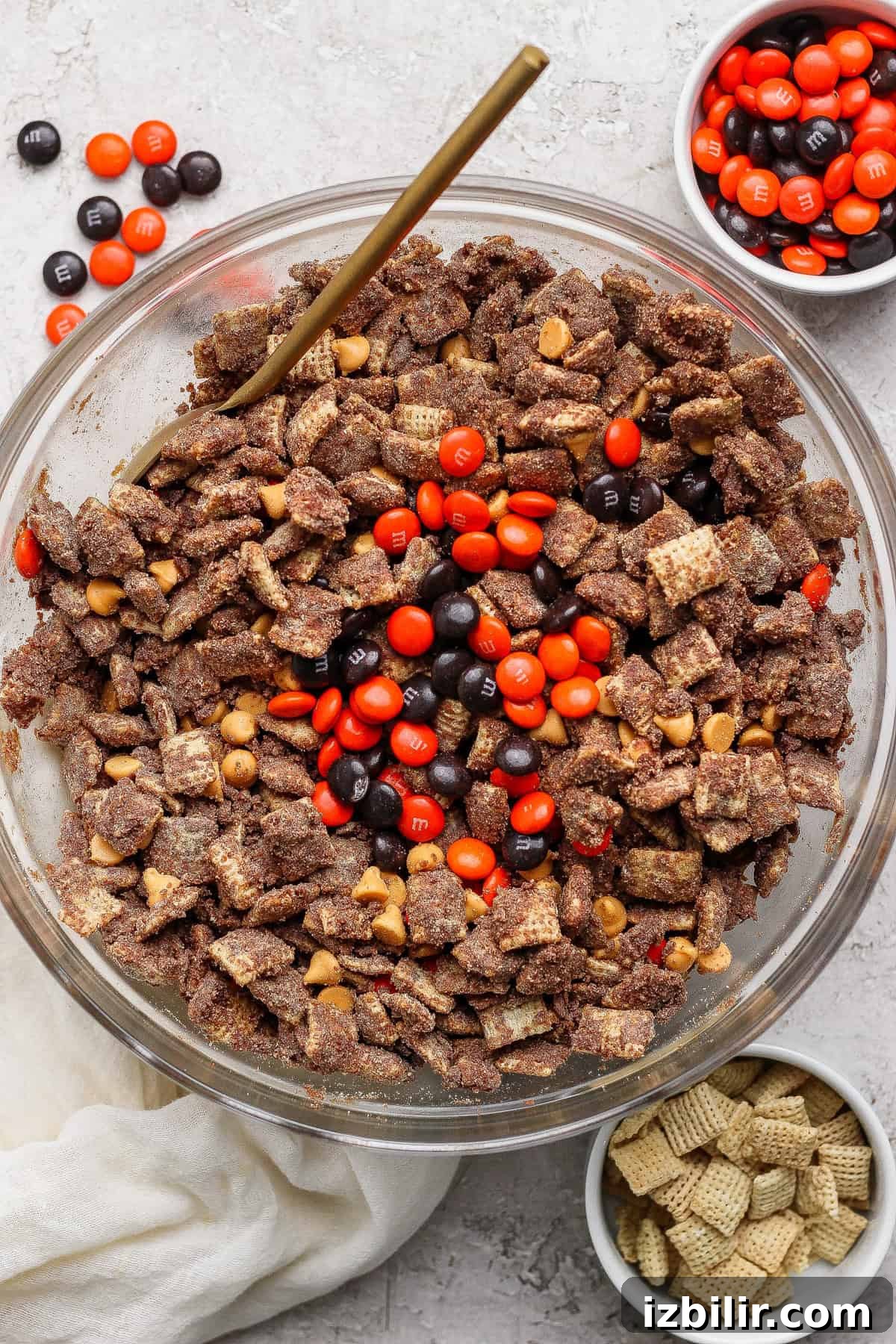 No-Bake Pumpkin Spice Protein Puppy Chow Recipe 2 A glass bowl filled with chocolate-coated Chex cereal, peanut butter chips, and orange and brown M&M candies; small bowls with extra M&Ms and plain Chex nearby.