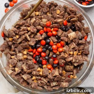No-Bake Pumpkin Spice Protein Puppy Chow Recipe 6 A glass bowl filled with chocolate-coated Chex cereal, peanut butter chips, and orange and brown M&M candies; small bowls with extra M&Ms and plain Chex nearby.